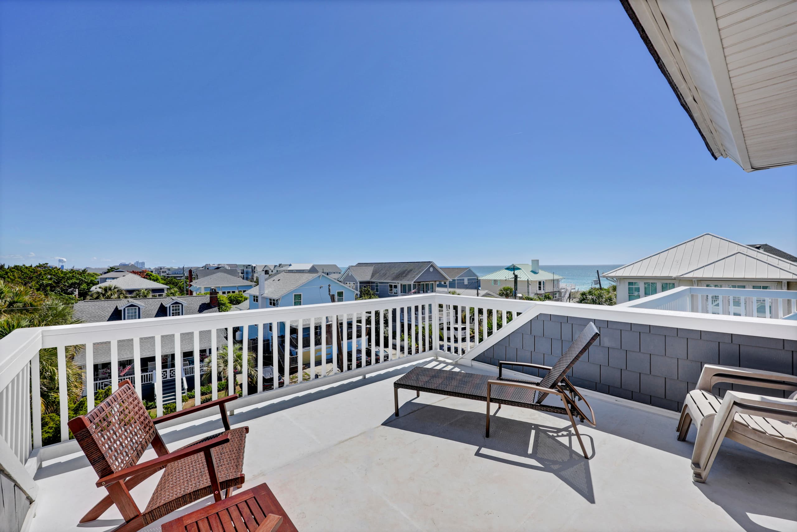 Elevated balcony offers coastal neighborhood views and nearby ocean glimpses. Sunbathe on loungers, relax in wicker chairs, and enjoy the bright Florida sky above.