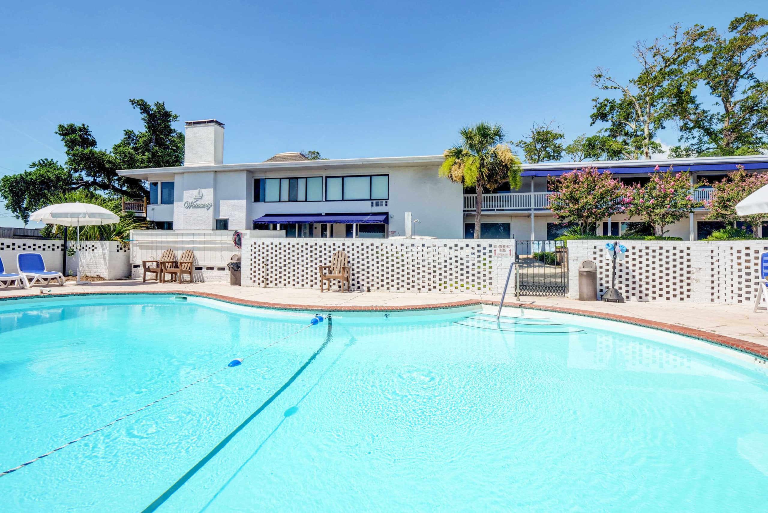 A refreshing outdoor community pool surrounded by breeze blocks, lounge chairs, and palm trees under clear blue skies. This property offers a peaceful poolside setting for sunny days.